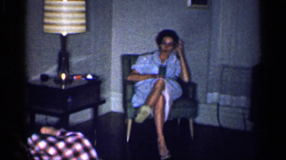 A woman in a light blue dress sits in a chair in the same Michigan living room, with a lamp, fireplace, and plaid fabric nearby.