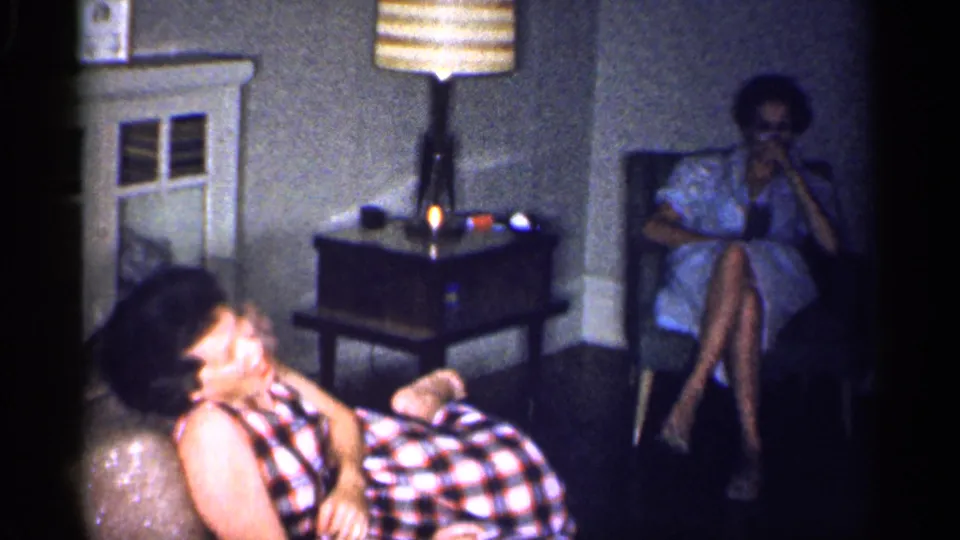 1959 Michigan home-movie still of one woman laughing in the foreground and another seated beside a lit table lamp.