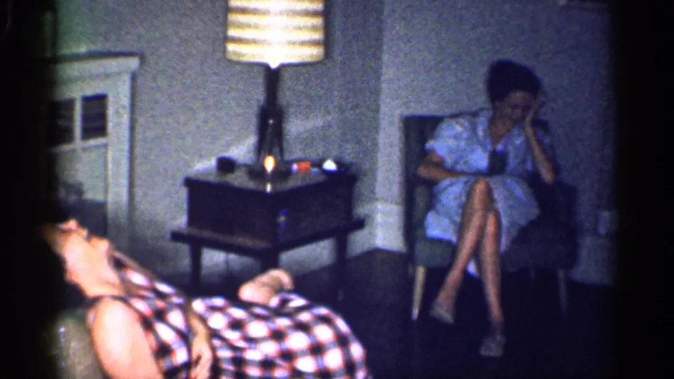 1959 Michigan living-room frame with a reclining laughing woman, a seated woman holding a glass, and a lit striped lamp.