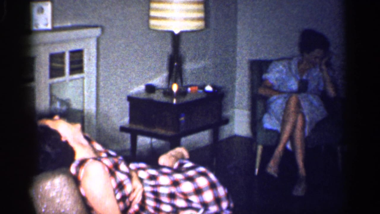 1959 Michigan home-movie frame showing two adult women in a dim mid-century living room with a couch, chair, lamp, and side table.