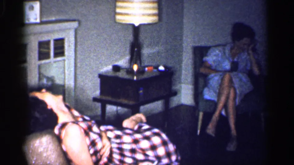 1959 Michigan home-movie frame showing two adult women in a dim mid-century living room with a couch, chair, lamp, and side table.