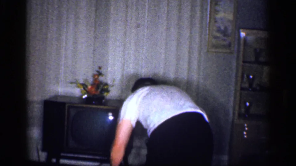 1959 Michigan home-movie frame showing an adult man bending toward a cabinet television with a small floral arrangement on top.