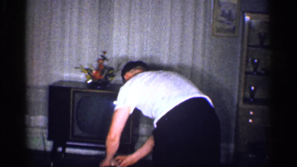 1959 Michigan home-movie frame showing a man leaning over a cabinet television in a wallpapered family living room.