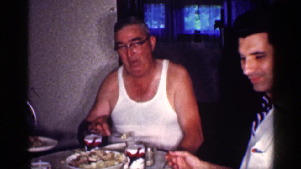 1959 Michigan home-movie frame of an older man in an undershirt seated beside another man at a table with plates and glasses.