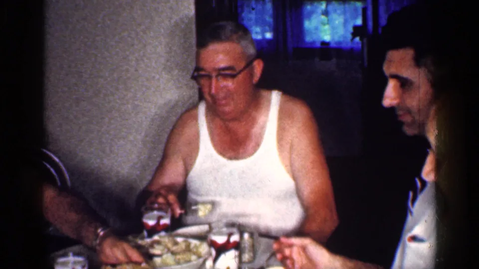 Adult men remain seated at a Michigan dining table, with food, glasses, and dark curtains visible in the 8mm frame.