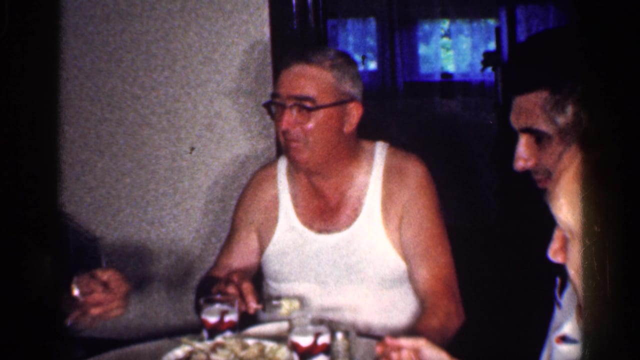 1959 Michigan home-movie frame showing adults gathered at a dining table, including a man in glasses and a white undershirt.