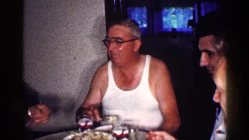 1959 Michigan home-movie frame showing adults gathered at a dining table, including a man in glasses and a white undershirt.
