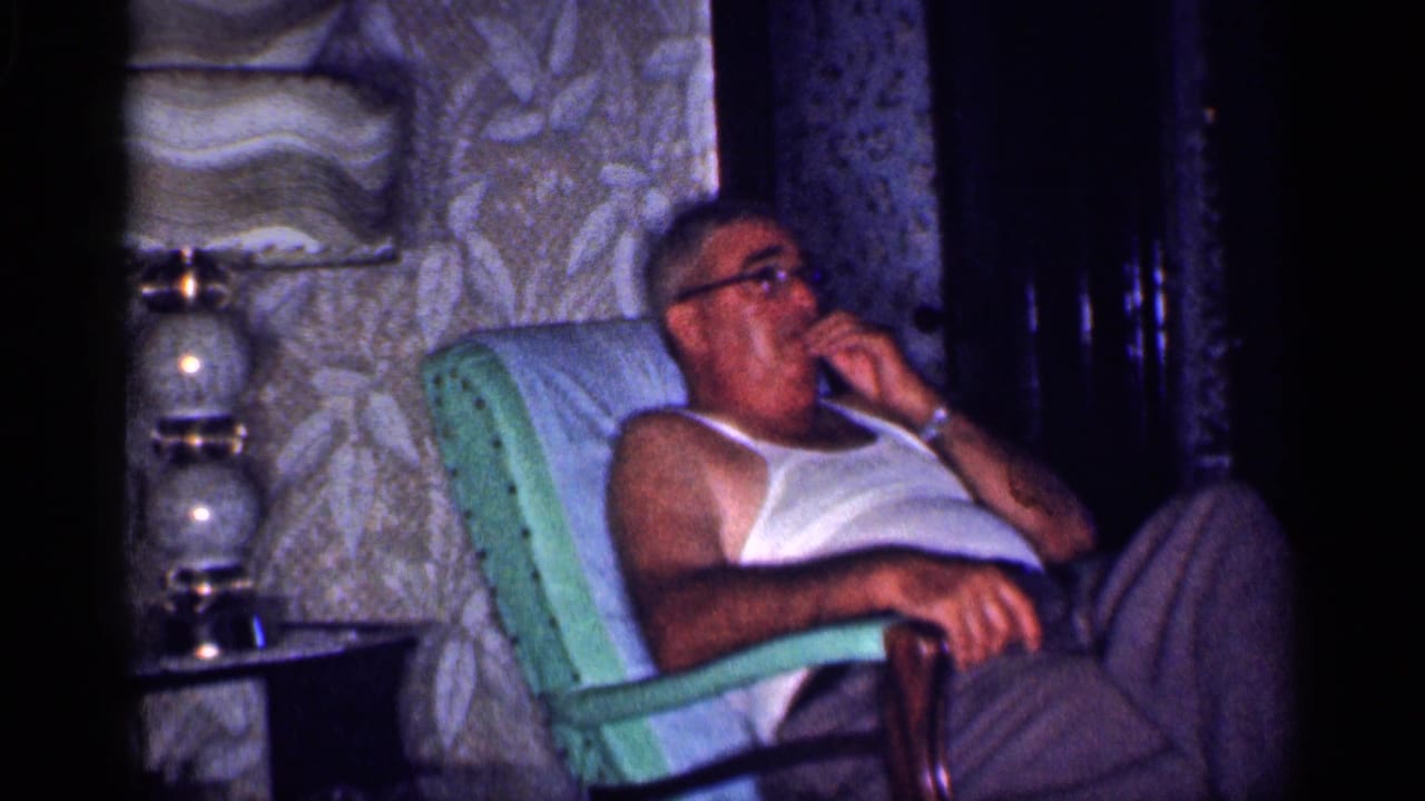 Man in glasses reclines in a green armchair beside patterned wallpaper and a glass lamp.