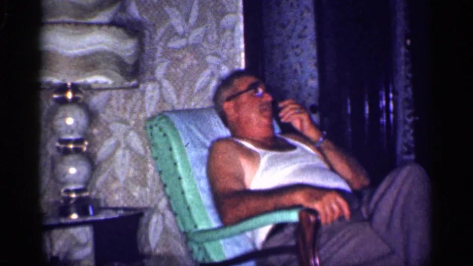 Home-movie frame from 1959 Michigan showing a man resting in a green armchair with a glass lamp nearby.