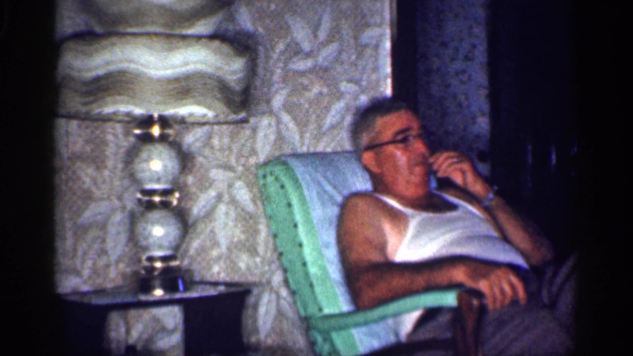 Man in a white tank top sits in a green chair using a telephone beside a lamp and floral wallpaper.