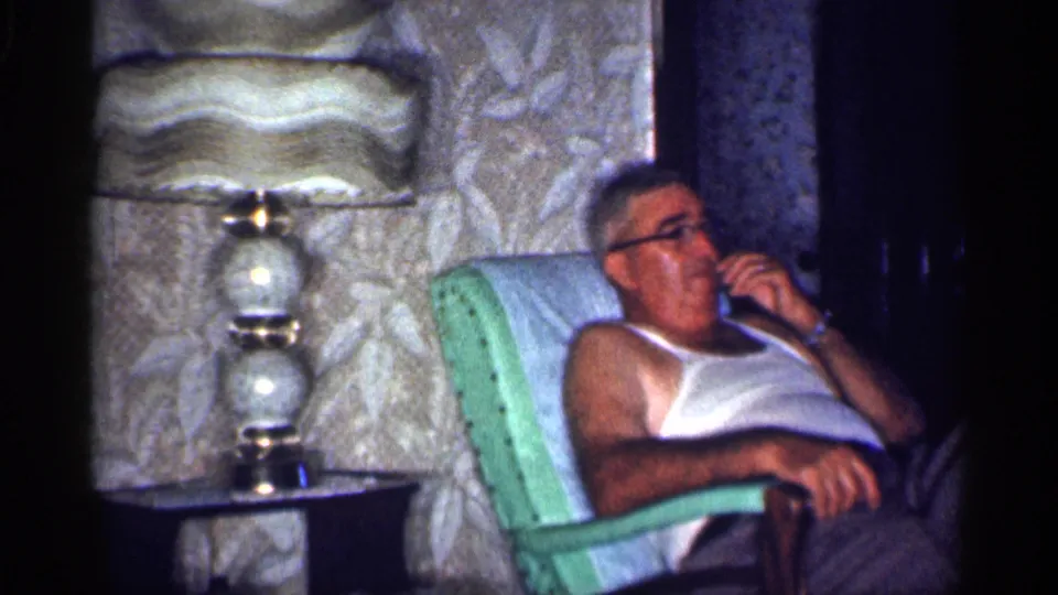 1959 Michigan home-movie frame of an adult man in a white tank top sitting in a green chair and using a telephone in a living room.