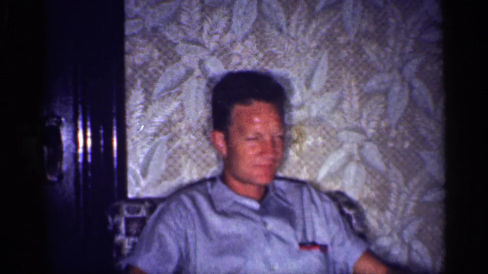 1959 Michigan interior frame showing an adult man in a light blue short-sleeve shirt seated in a vintage living room.