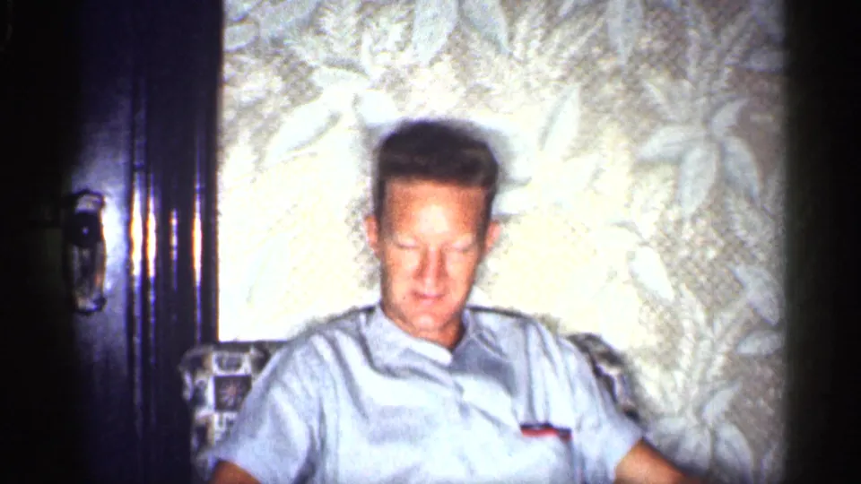 1959 Michigan home interior frame of a seated adult man in a light shirt, softly lit against floral wallpaper.