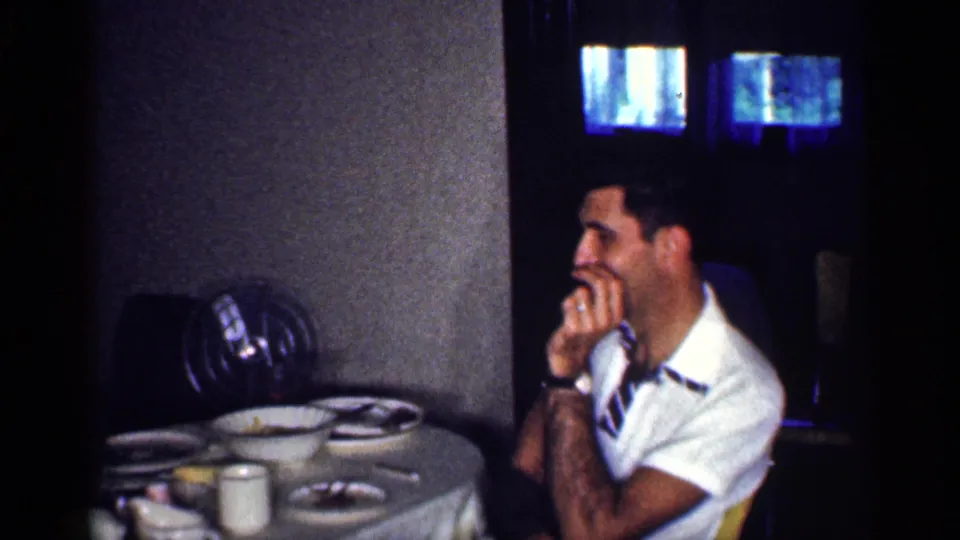 1959 Michigan home-movie frame of a smiling adult man at a dining table with dishes, a striped tie, and a small television nearby.