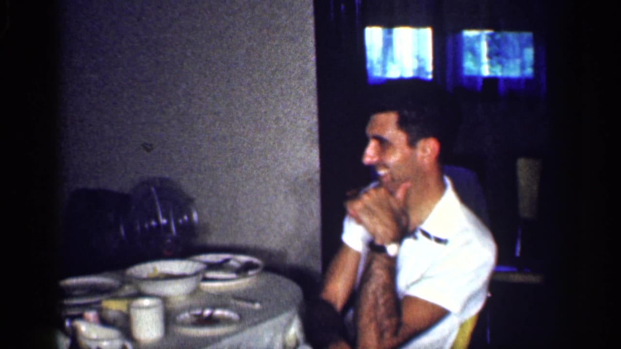 Man in a white shirt laughs beside a table set with dishes and cups.
