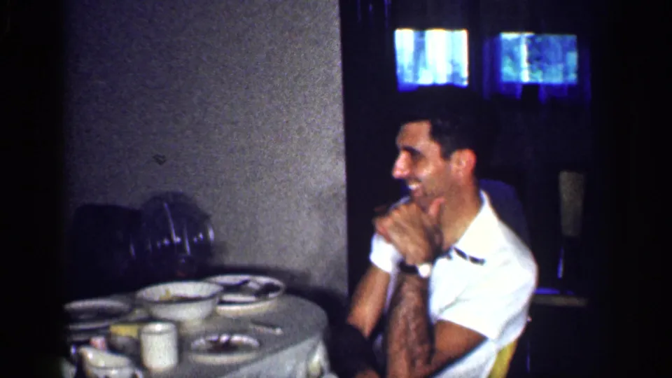 In a 1959 Michigan home interior, a man in a white shirt laughs at a dinner table with dishes, cups, and an oscillating fan nearby.