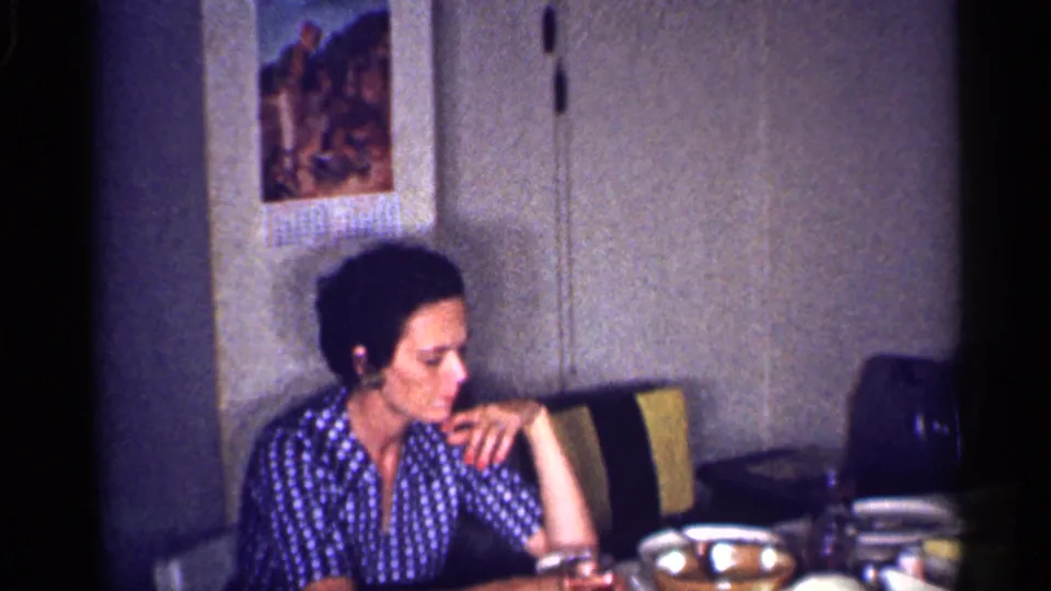 The woman remains seated in the 1959 Michigan dinner-table scene, looking down as her hand rests near her face amid dishes and cups.