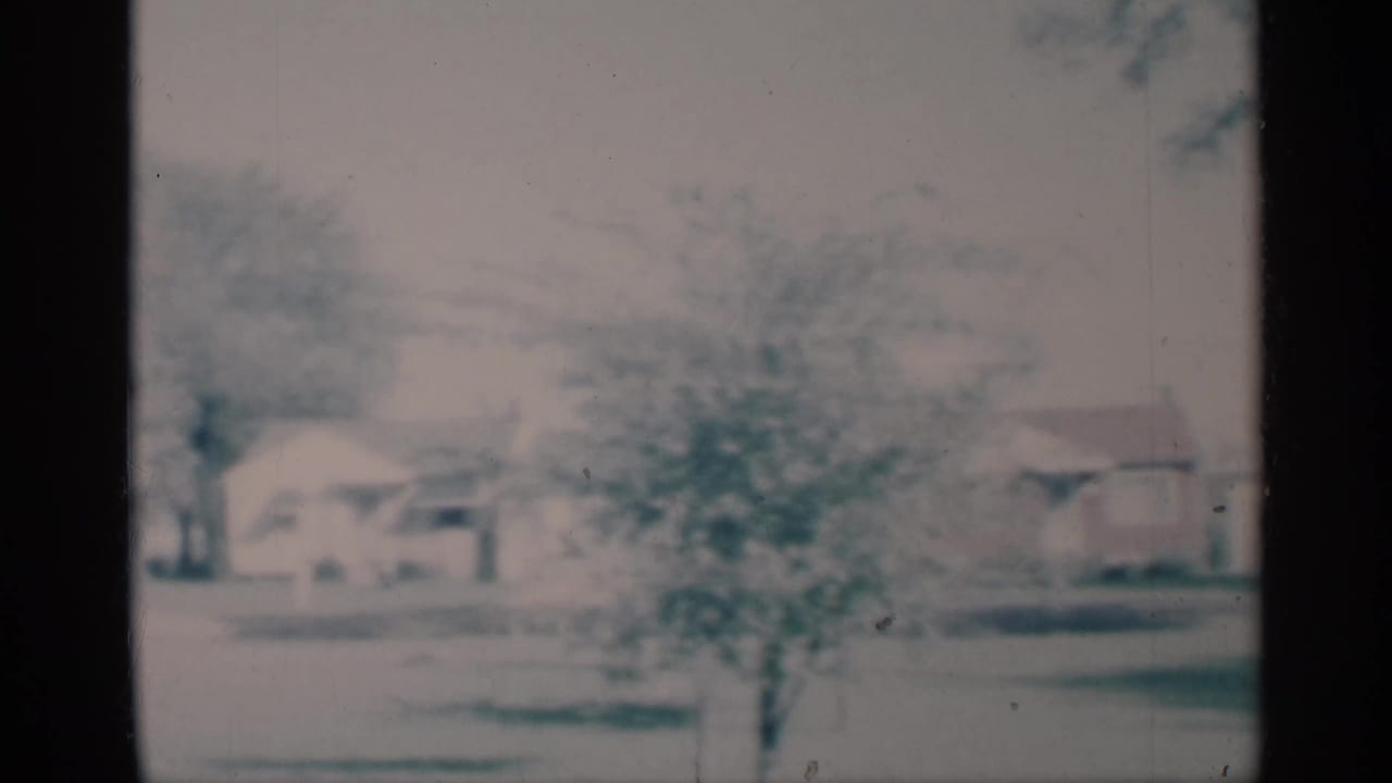 Soft 8mm view of a snowy suburban street with houses, trees, and a driveway.