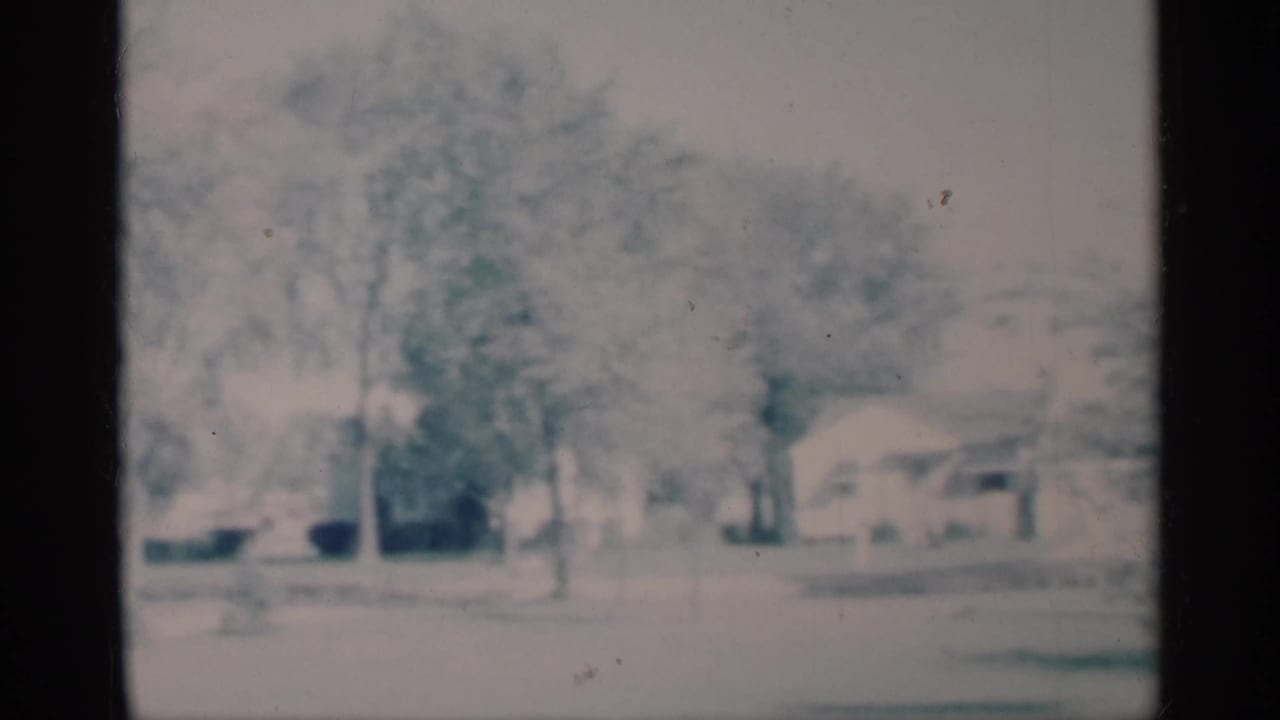 Snowy residential view with trees, houses, and a parked vehicle partly visible.