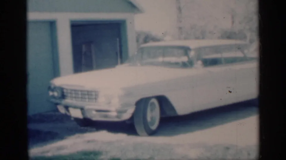 1959 Michigan home-movie frame of a white mid-century sedan with whitewall tires parked beside a garage in snow.
