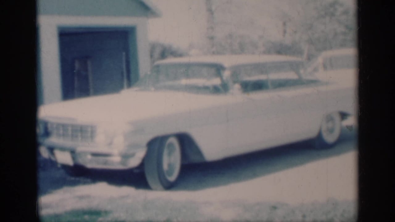 1959 Michigan 8mm frame showing a white midcentury sedan with chrome trim parked outside a suburban house.