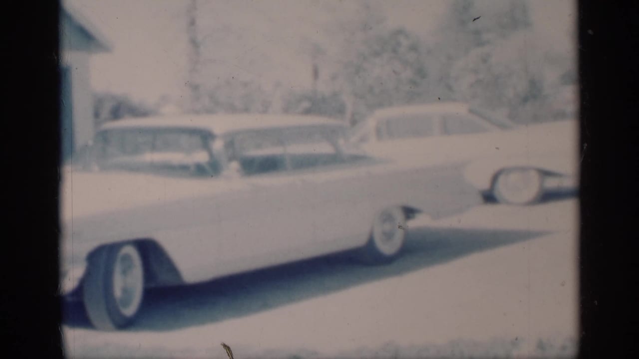 Soft 8mm view of a white late-1950s sedan in a driveway, with additional parked cars visible in the background.