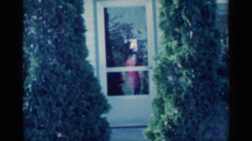 1959 Michigan home-movie frame looking toward a glass door between shrubs, with a person in red visible inside the house.