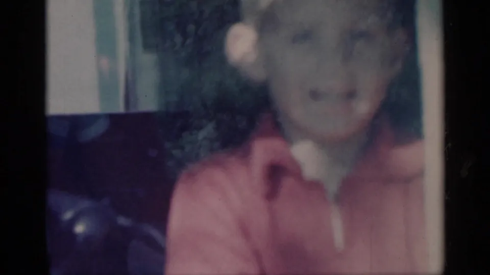 A soft 1959 Michigan home-movie frame shows a blurred young boy in a red top near a shaded yard or exterior threshold.
