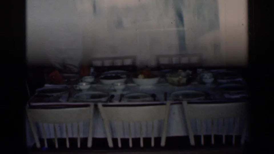 Interior home-movie frame showing a dining table prepared with plates, bowls, chairs, and food in a private household setting.