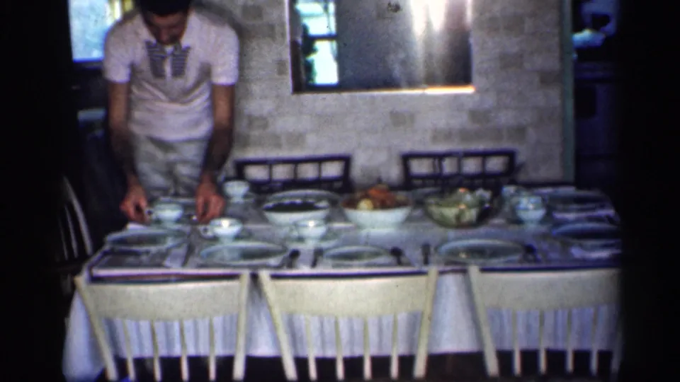 1959 Michigan interior frame showing a man arranging dishes at a set dining table in a private home.