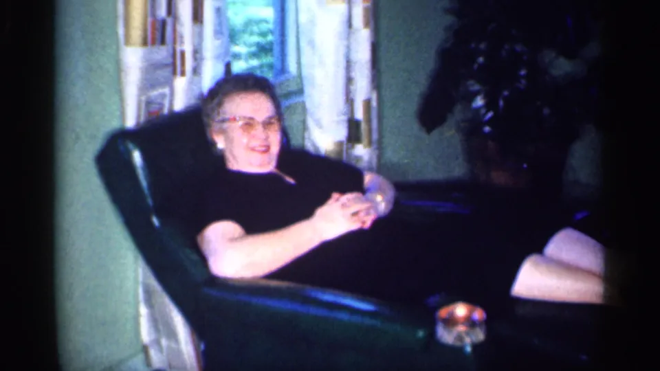 1959 Michigan home-movie frame of an adult woman in glasses smiling from a green armchair in a private interior.