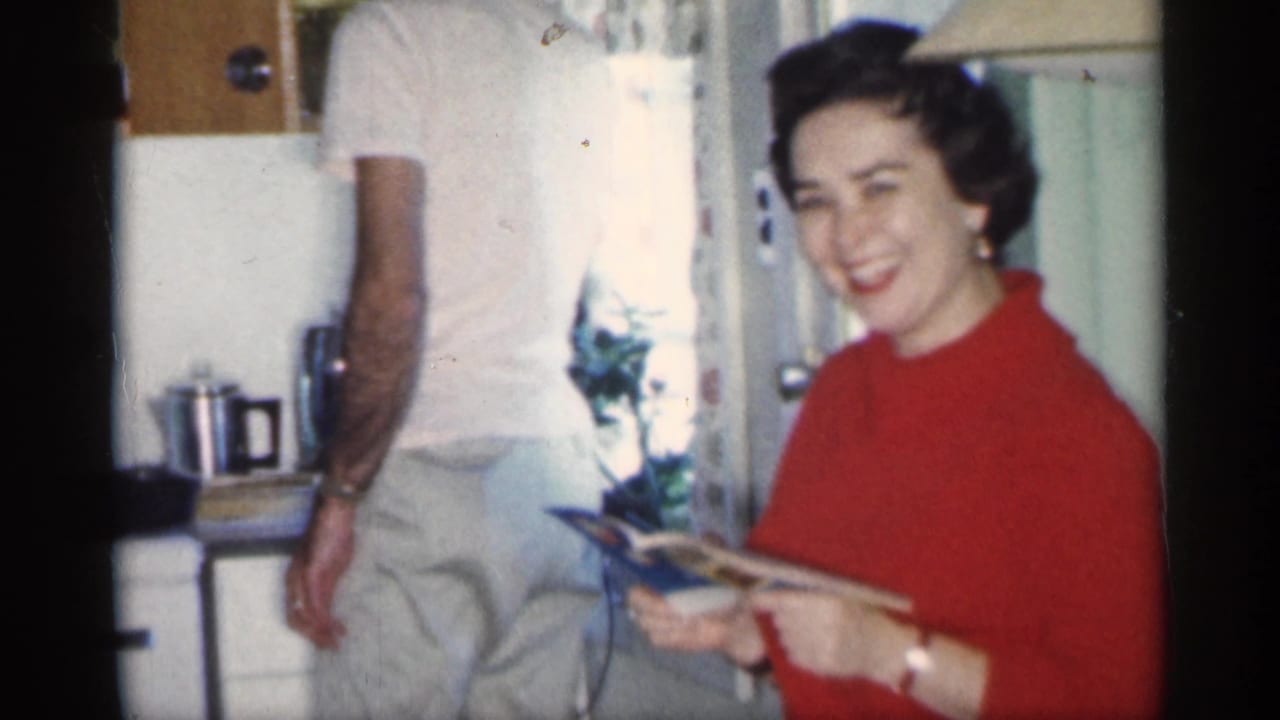 Woman in a red sweater smiles in a kitchen while holding a magazine near a man in white.
