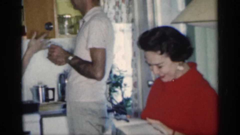 1959 Michigan home-movie frame of a woman looking down at a magazine in a kitchen with a man standing beside her.