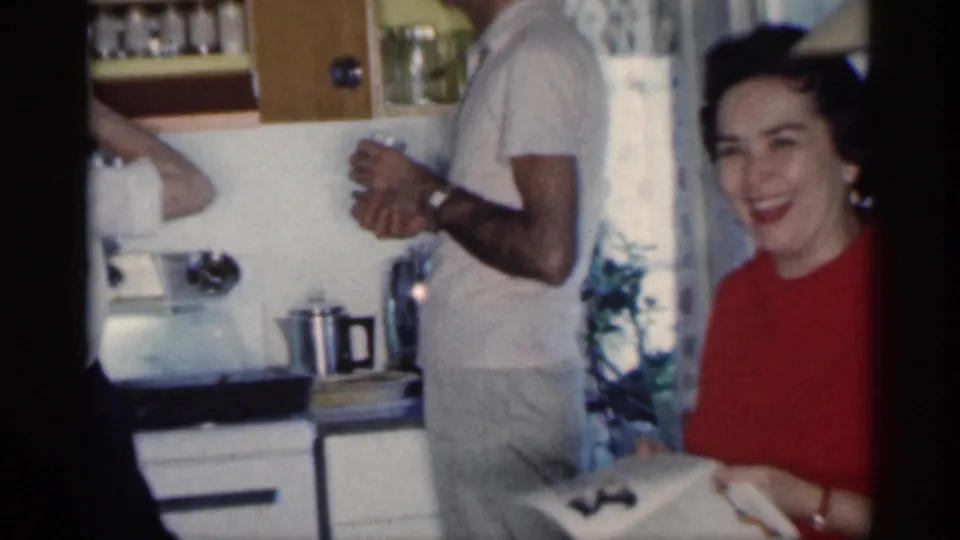1959 Michigan kitchen frame showing a woman laughing toward the camera with a man partly visible beside the counter.