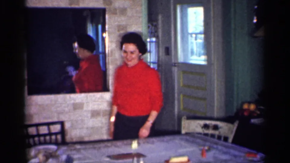 In a 1959 Michigan home interior, a woman in a red top stands near a covered dinner table with chairs, doors, and a wall mirror visible.