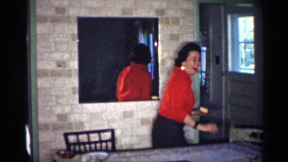 The woman in red moves past the dinner table in a Michigan home interior, with chairs, a mirror, and painted doors framing the scene.
