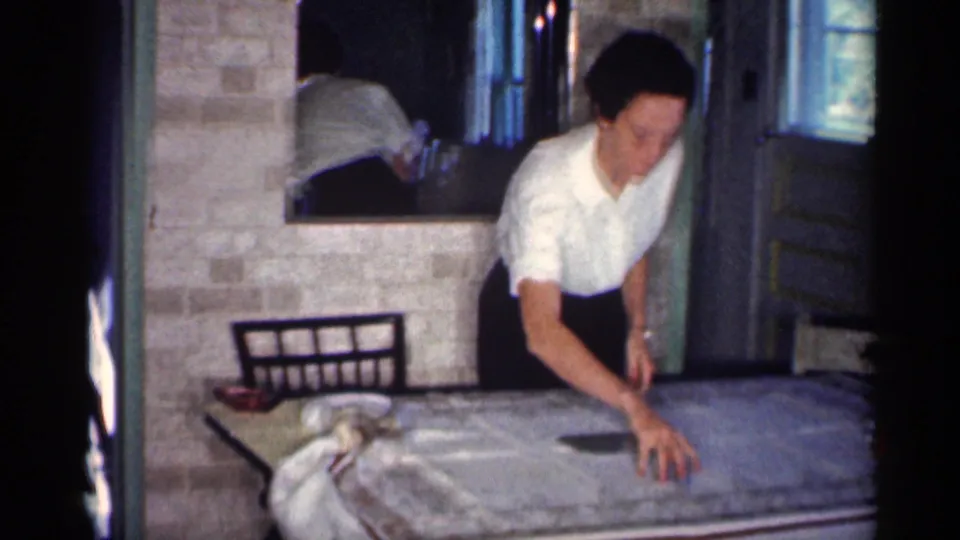 A woman in a white blouse leans across a dinner table in the same 1959 Michigan home interior, with tableware and a wall mirror behind her.