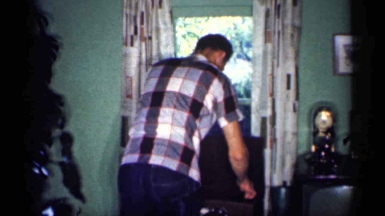 Man in a plaid shirt bends near a chair beside a window with patterned curtains.