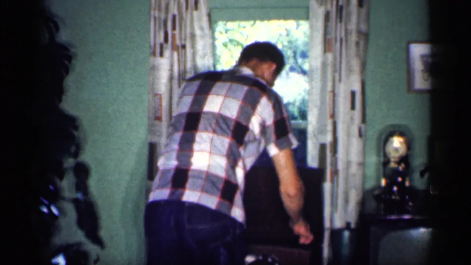 In the Michigan home-movie scene, a man in a plaid shirt adjusts or reaches toward a chair near a bright curtained window.