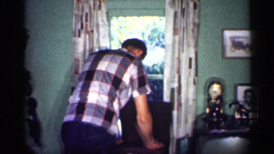 A man in a plaid shirt bends near a chair in a 1959 Michigan home interior, with curtains, a clock, and framed photographs visible nearby.