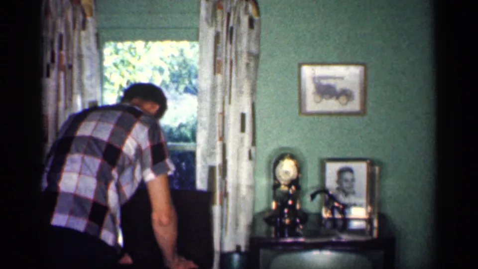 The man remains near the chair in the Michigan home interior, with a curtained window, green wall, clock, and personal photographs in view.