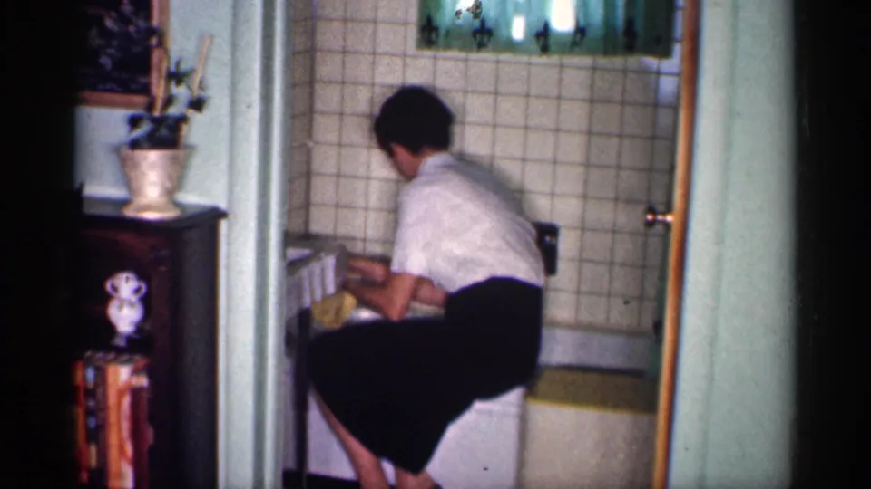The woman bends over the bathtub, using a yellow cloth near the tub edge in a tiled 1959 Michigan bathroom.