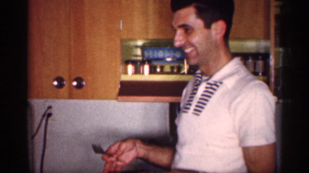 Smiling man in a white striped polo stands in a small kitchen near wood cabinets.