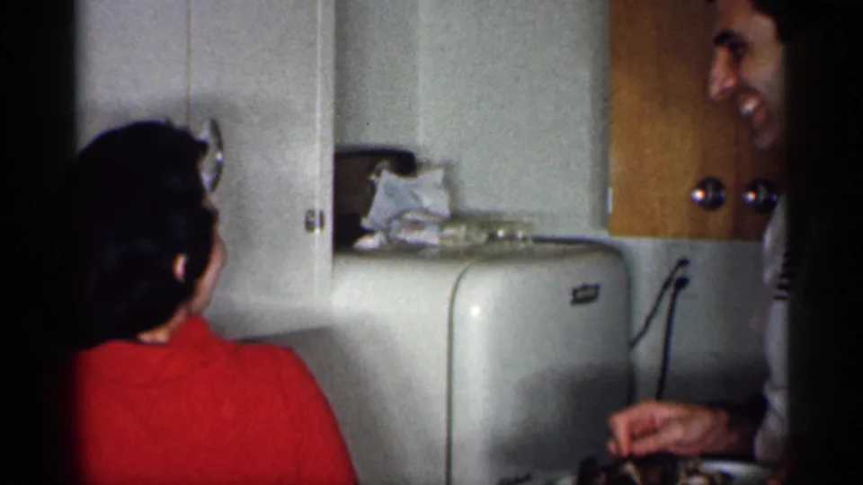 A woman in a red sweater faces away toward a kitchen wall while a man is partly visible at the edge of the frame.