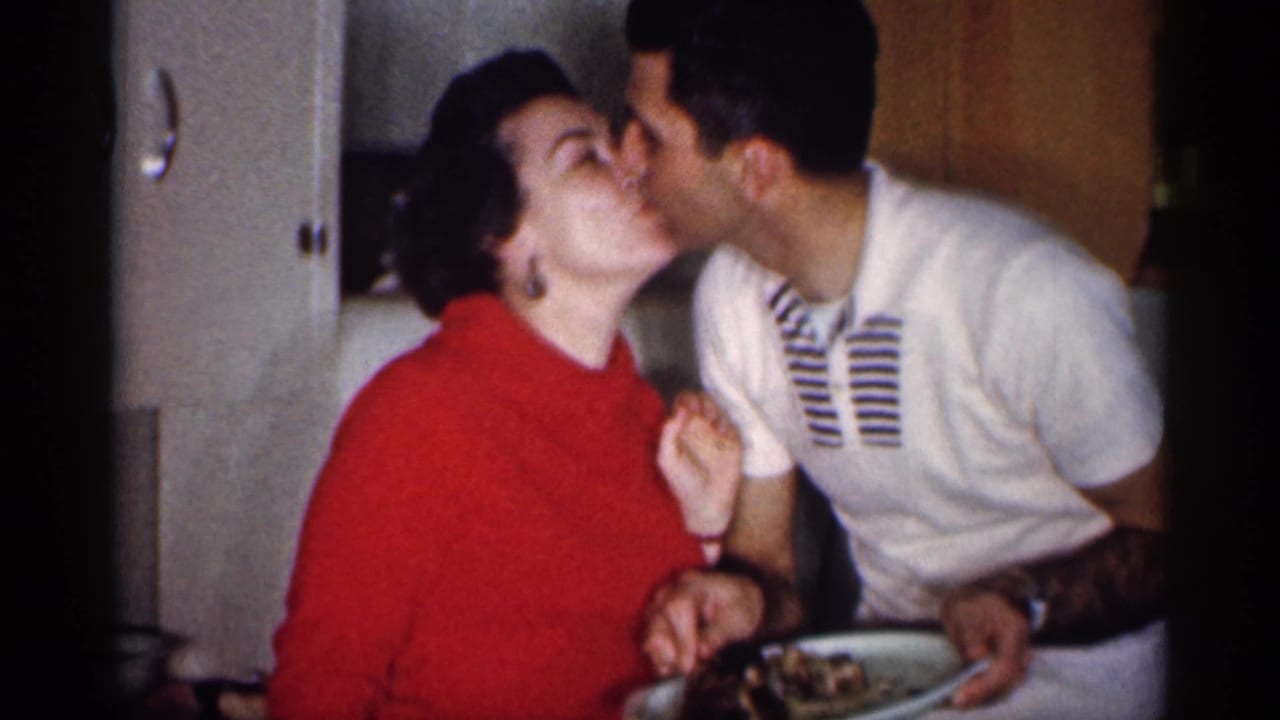 A woman in a red sweater kisses a man holding a plate in a kitchen.