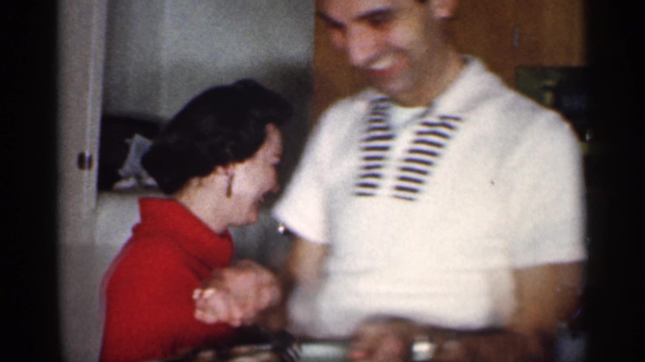A laughing couple stands close together in a kitchen with cabinets behind them.