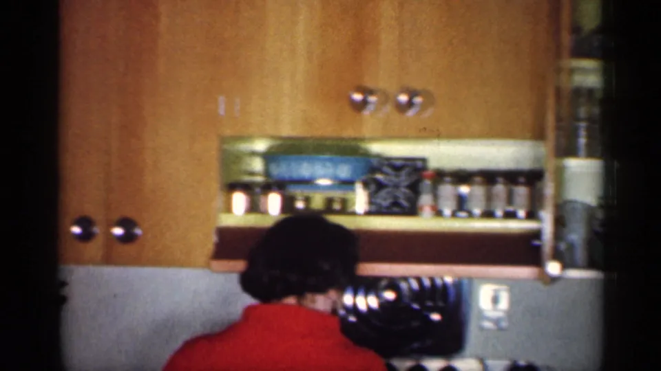 The woman in red turns toward a white electric stove below wood cabinets and shelves holding kitchen containers.