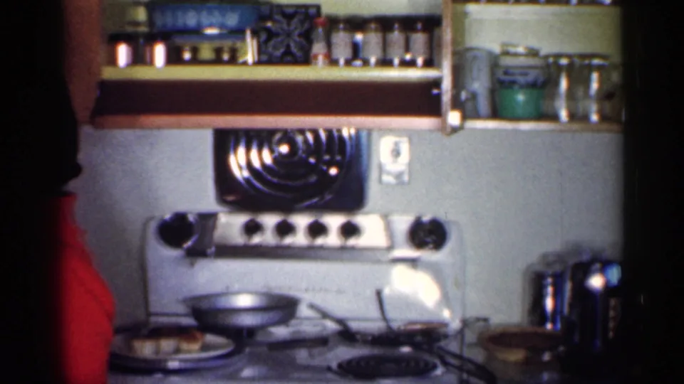 A white electric range with black burner details, pans, shelves, and kitchenware appears as the woman in red stands partly at the side.