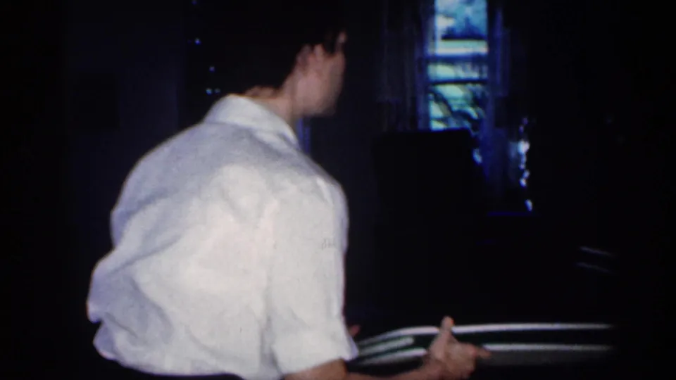 A man in a white shirt turns in a darker kitchen area, holding objects near a window or doorway.