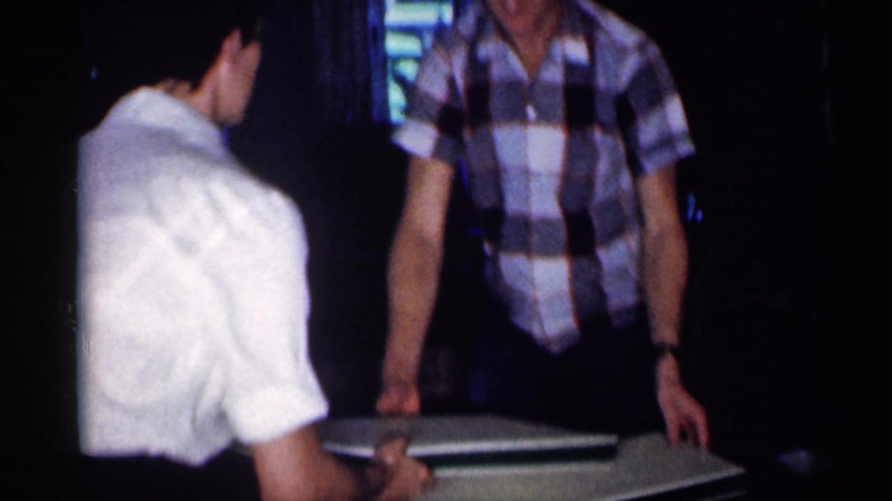 Two adults stand by a dining table with a white rectangular panel in a dim interior.